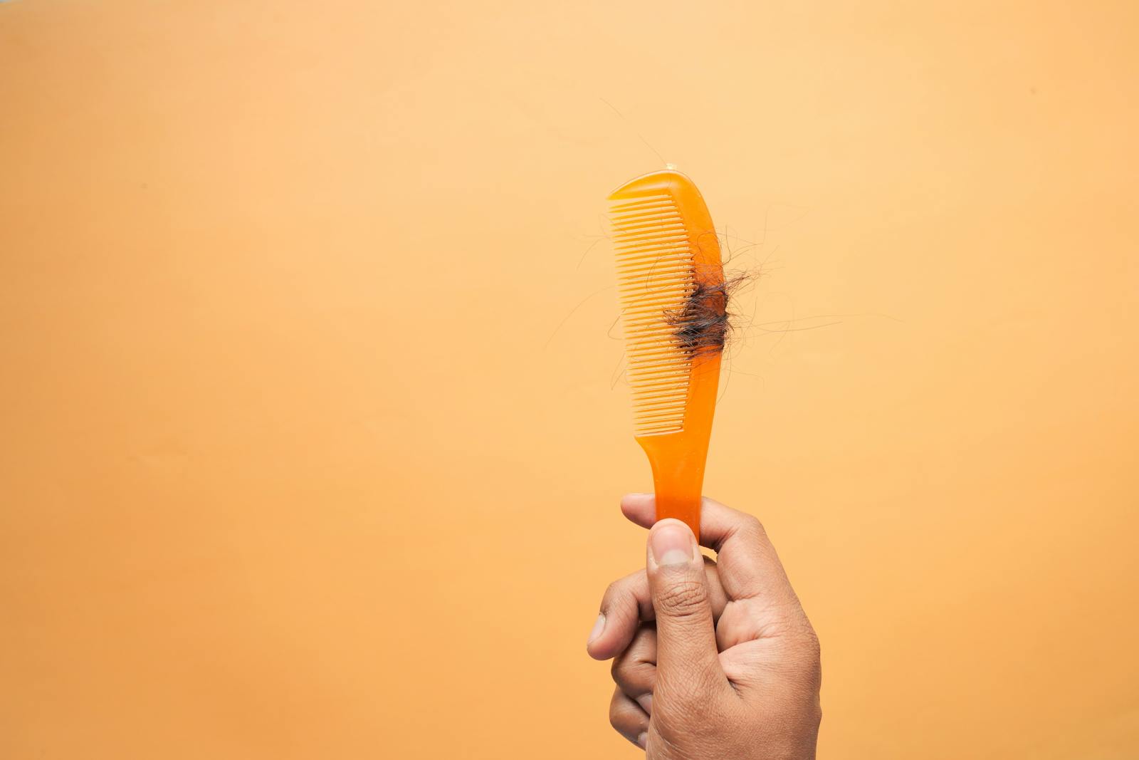 Close-up of hand holding a comb with hair against an orange backdrop, highlighting hair loss concept.