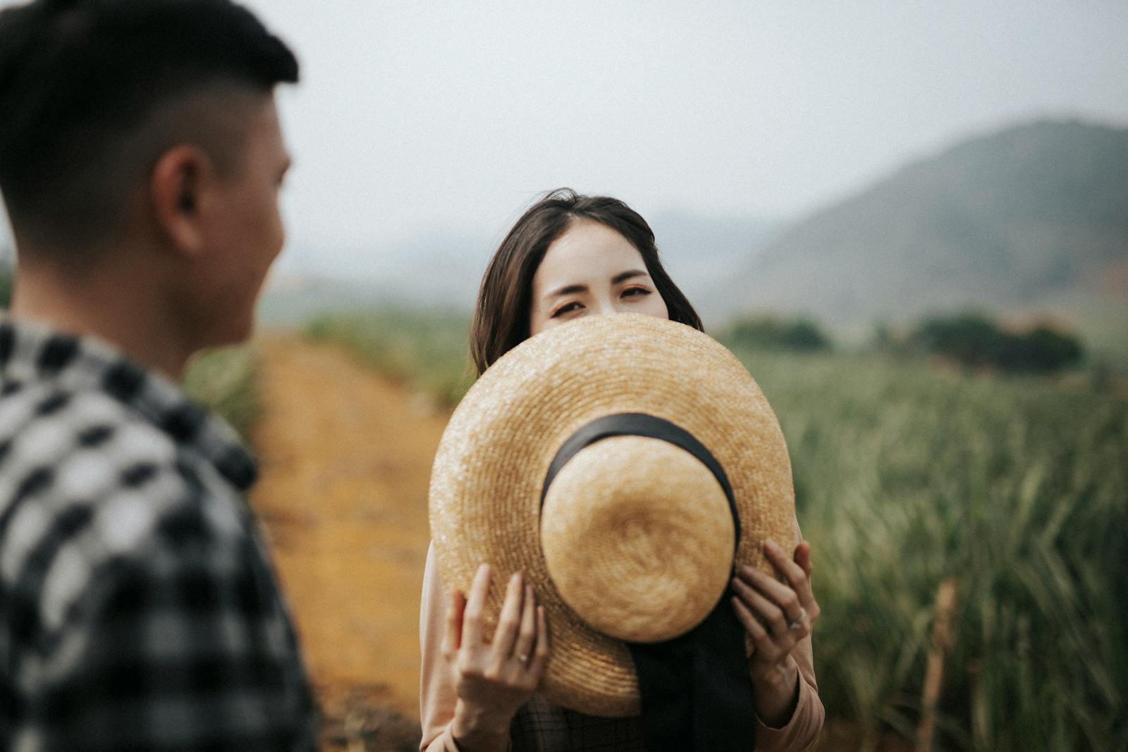 A woman playfully hides behind a straw hat in a Vietnamese field. Natural and candid moment.