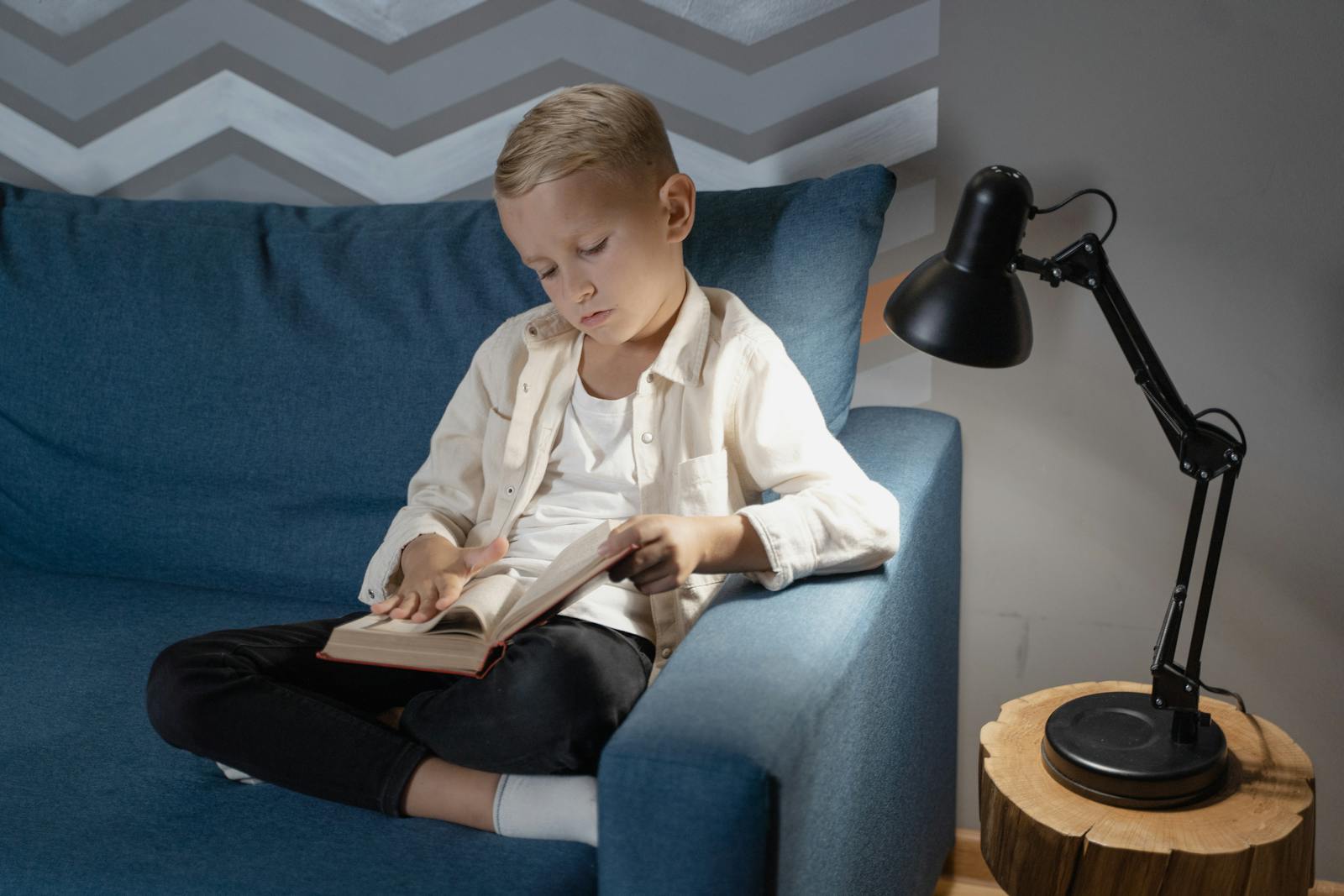 A young boy attentively reading a book while sitting on a cozy blue couch indoors next to a lamp.