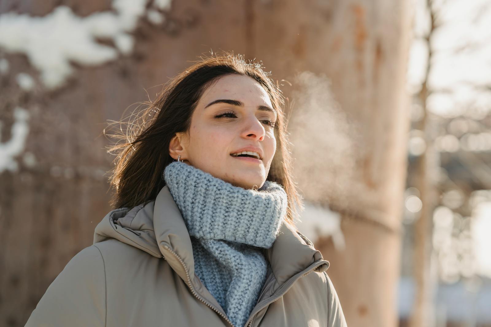 A serene young woman in warm clothing enjoys a sunny winter day outdoors, exhaling visible breath.
