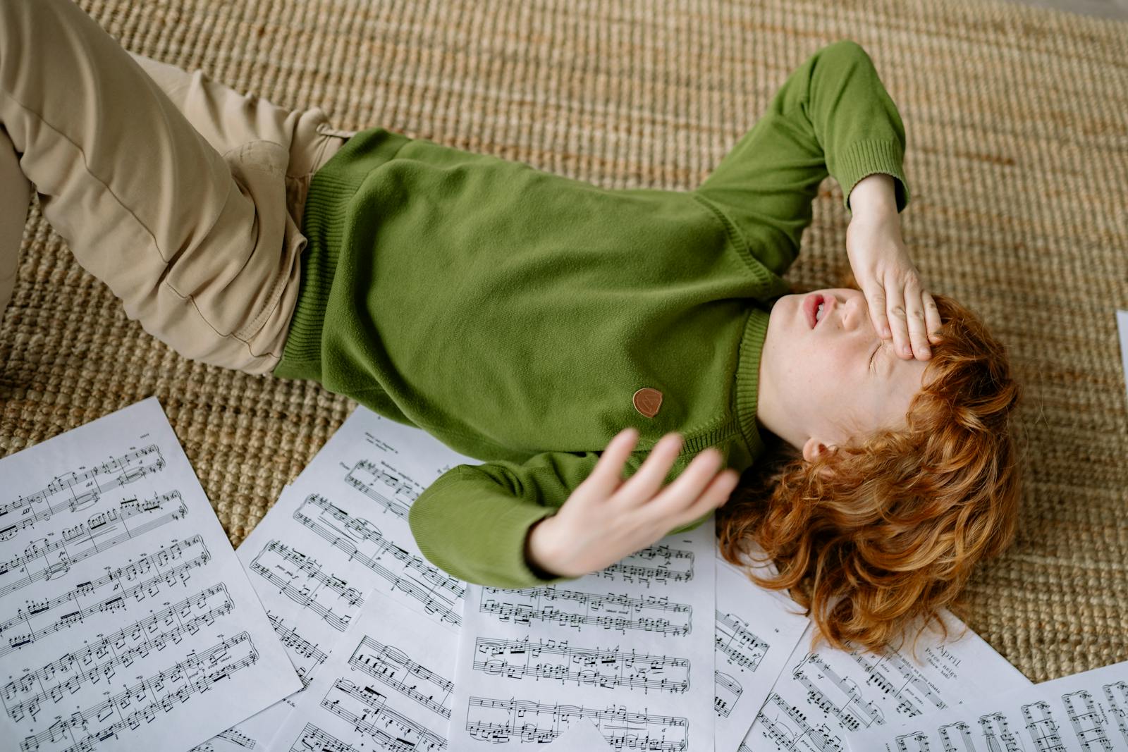 A child lying on a rug surrounded by music sheets, expressing frustration.